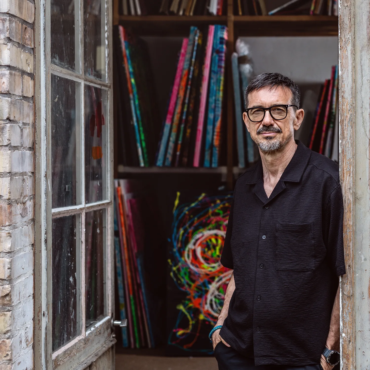 The artist standing in the open studio doorway, with shelves of stacked canvases and a colourful work behind him