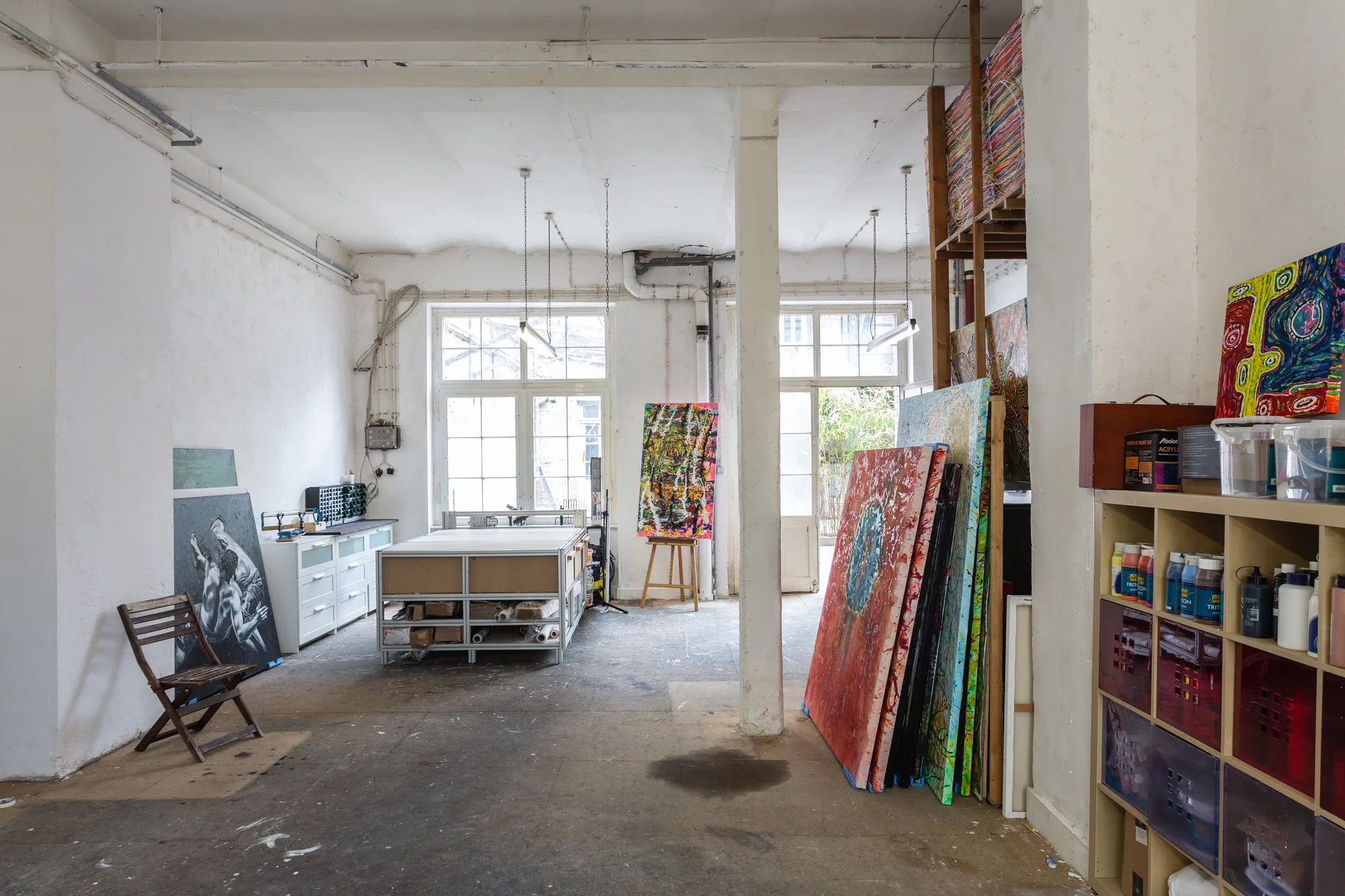 A look into the studio: a bright room with a work table, easel, a shelf of paints and canvases leaning against the wall