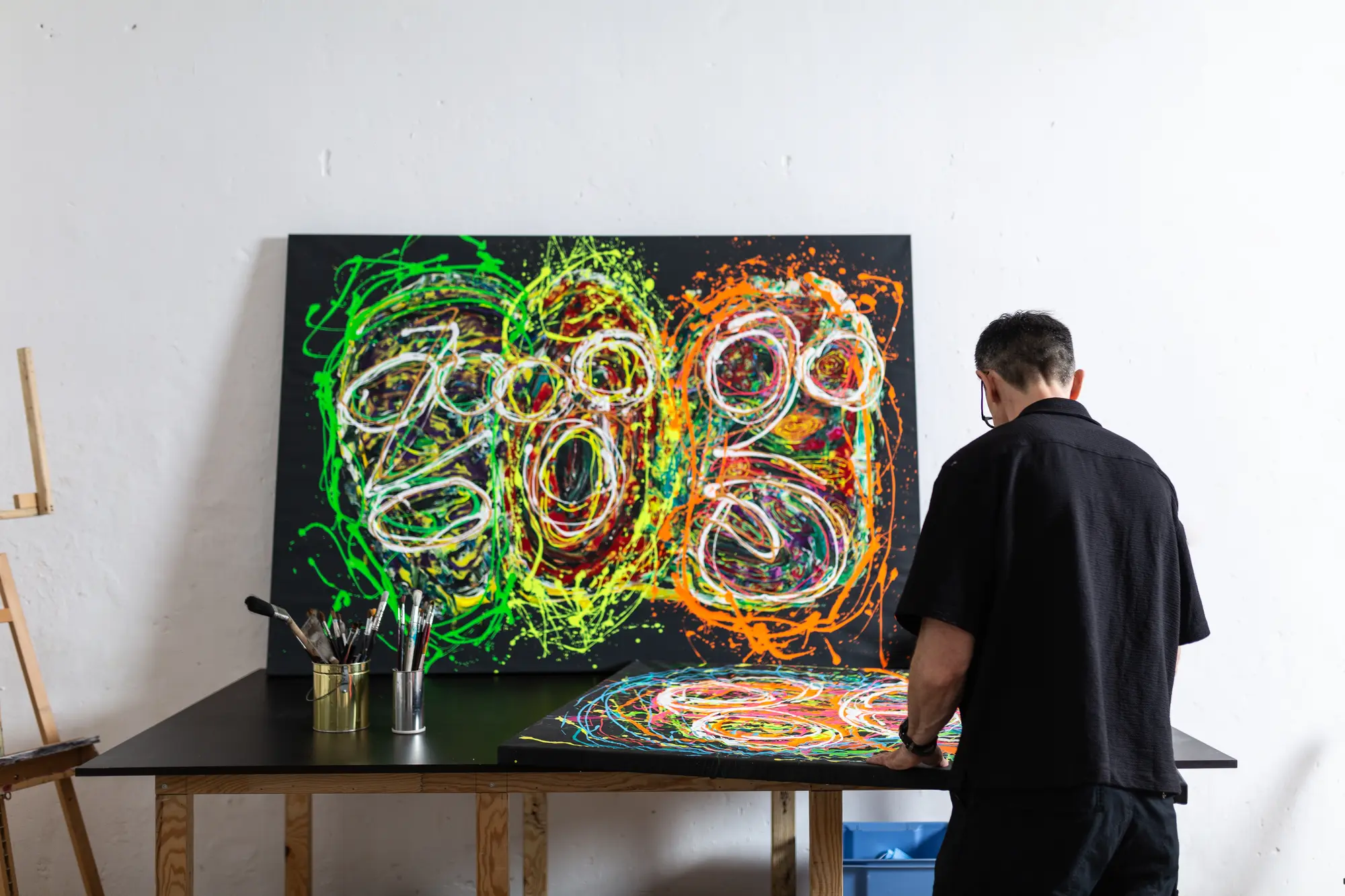 The artist working at the studio table on a colourful piece, with a large-format painting of green and orange looping lines behind him